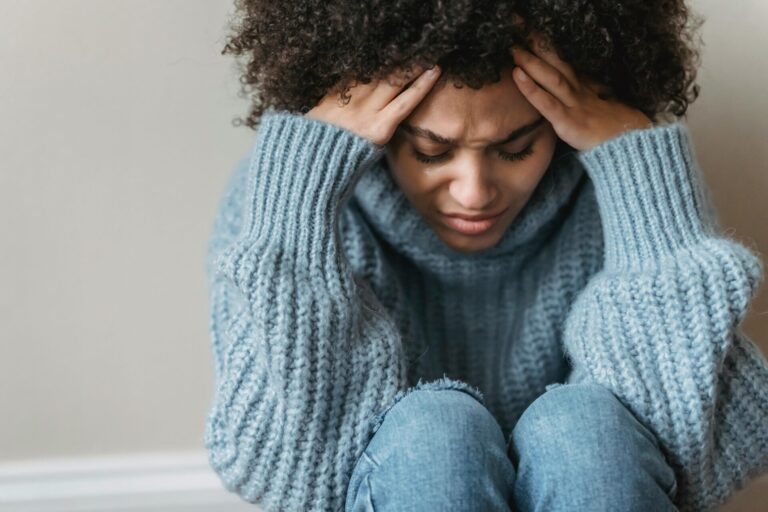 Image of a woman in blue sweater holding head in distress, sitting with eyes closed, showing how to handle seasonal depression in Huntsville winters.