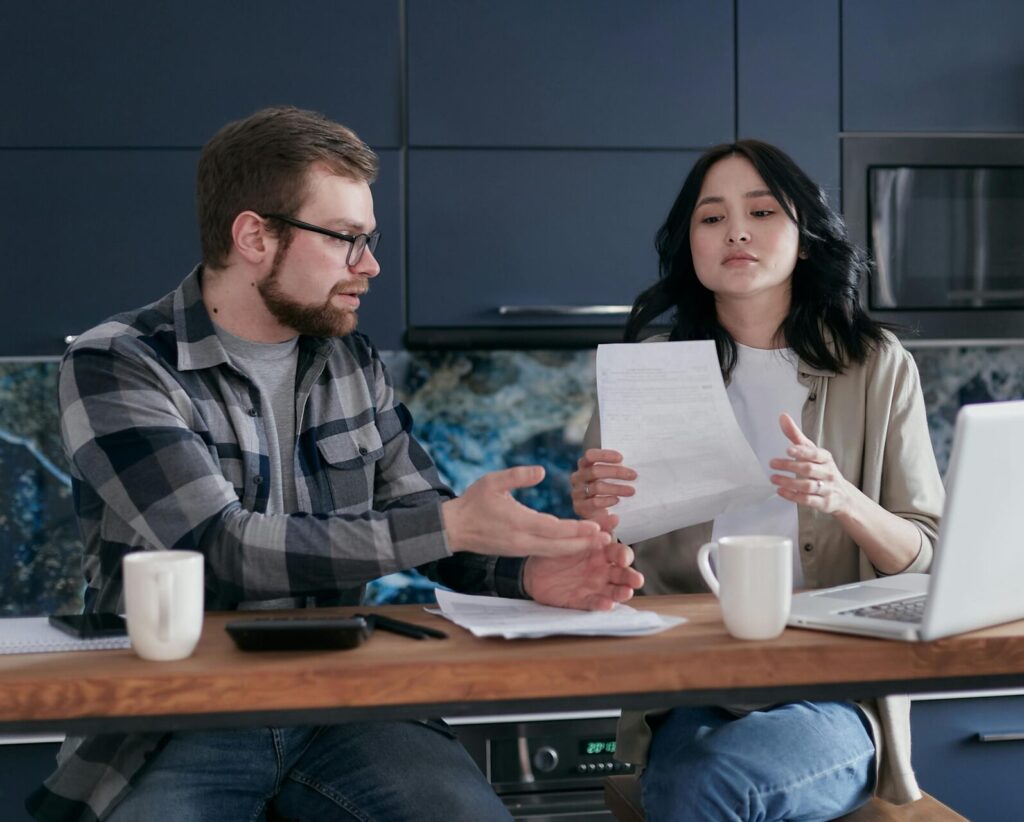 Image of a couple sits at a table discussing finances, with papers, a laptop, and coffee mugs in front of them.