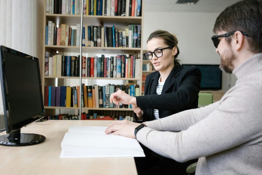 Image of a woman helps a blind man read Braille in a library, with bookshelves and a computer in the background.