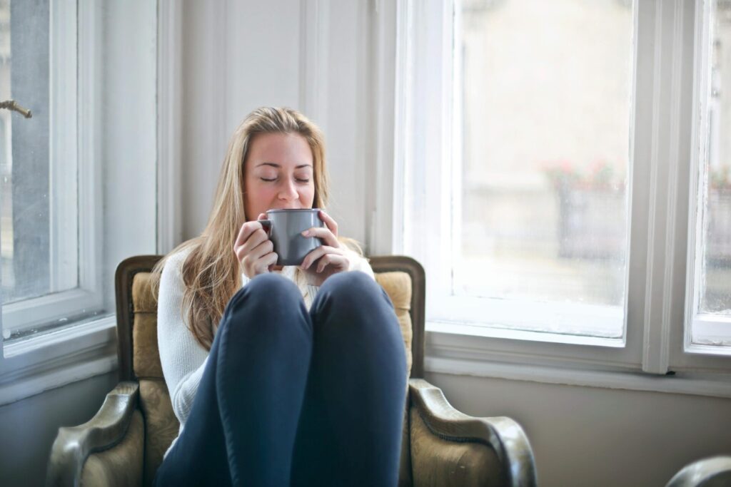 Image of a woman sitting by window with knees up, holding mug and smiling peacefully.