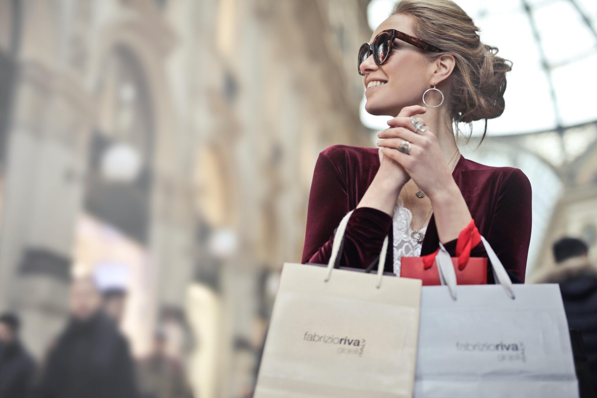 Image of a smiling woman in sunglasses holding shopping bags, enjoying a day out in a stylish urban setting.