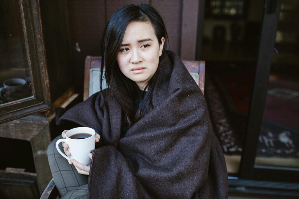 Image of a woman wrapped in blanket holding mug, looking uneasy while sitting outside.