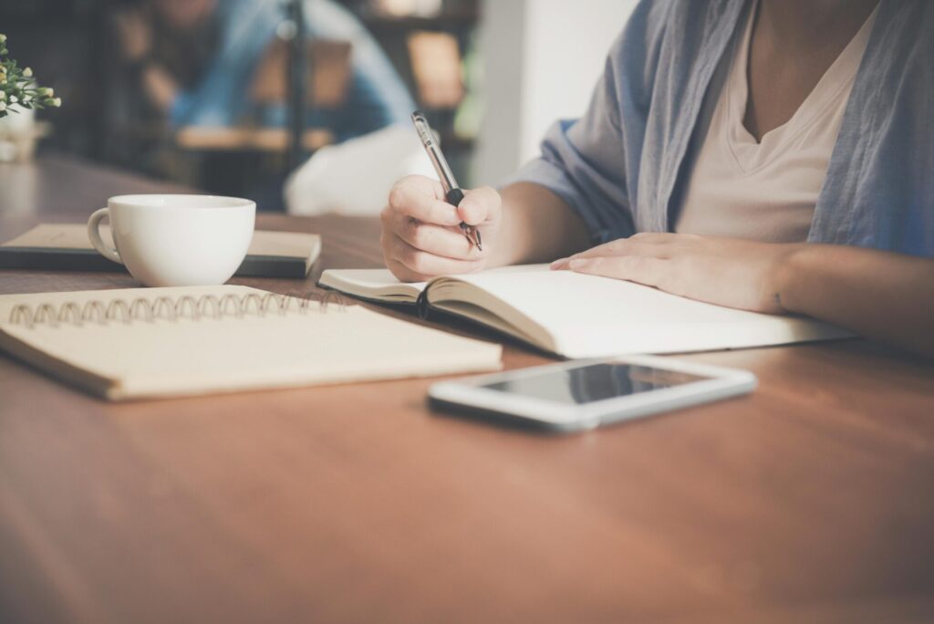 Image of a women writing down on a diary on table with a white cup of coffee and mobile phone on it, representing  Huntsville moms.