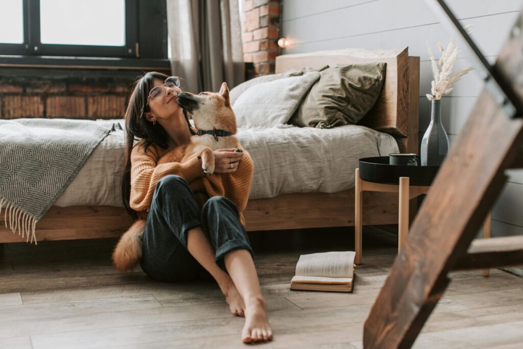 Image of a smiling woman in glasses hugs and plays with her dog while sitting on the floor beside her bed.
