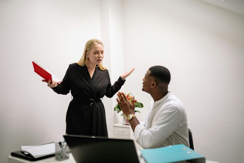 Image of a oman standing with a folder arguing with man at desk in a modern white office setting.