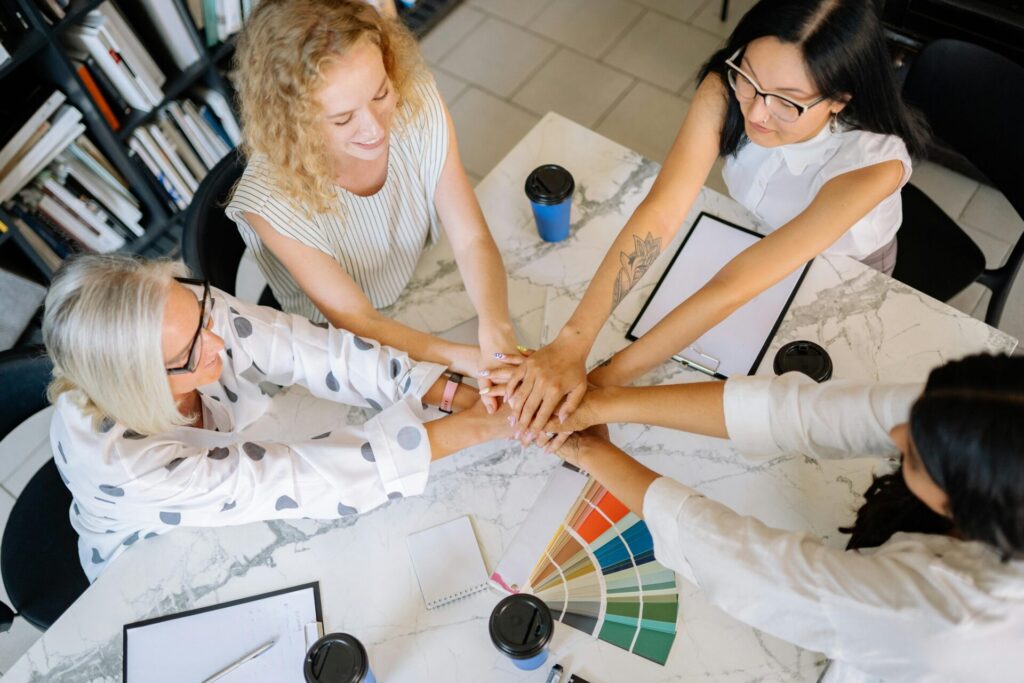 Image of a team members placing their hands together over a table with notebooks, coffee cups, and color samples.